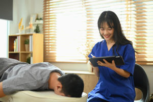 Smiling female back pain specialist using a digital tablet while examining male patient on the bed at clinic.