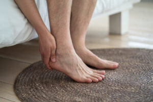 A man is suffering from foot and heel pain while holding his heel in his bedroom.
