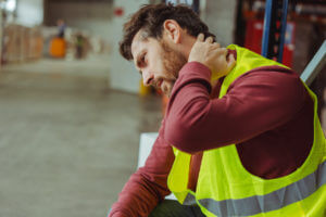 A tired, upset, sick, bearded worker experiencing neck and back pain after a shift in the warehouse.